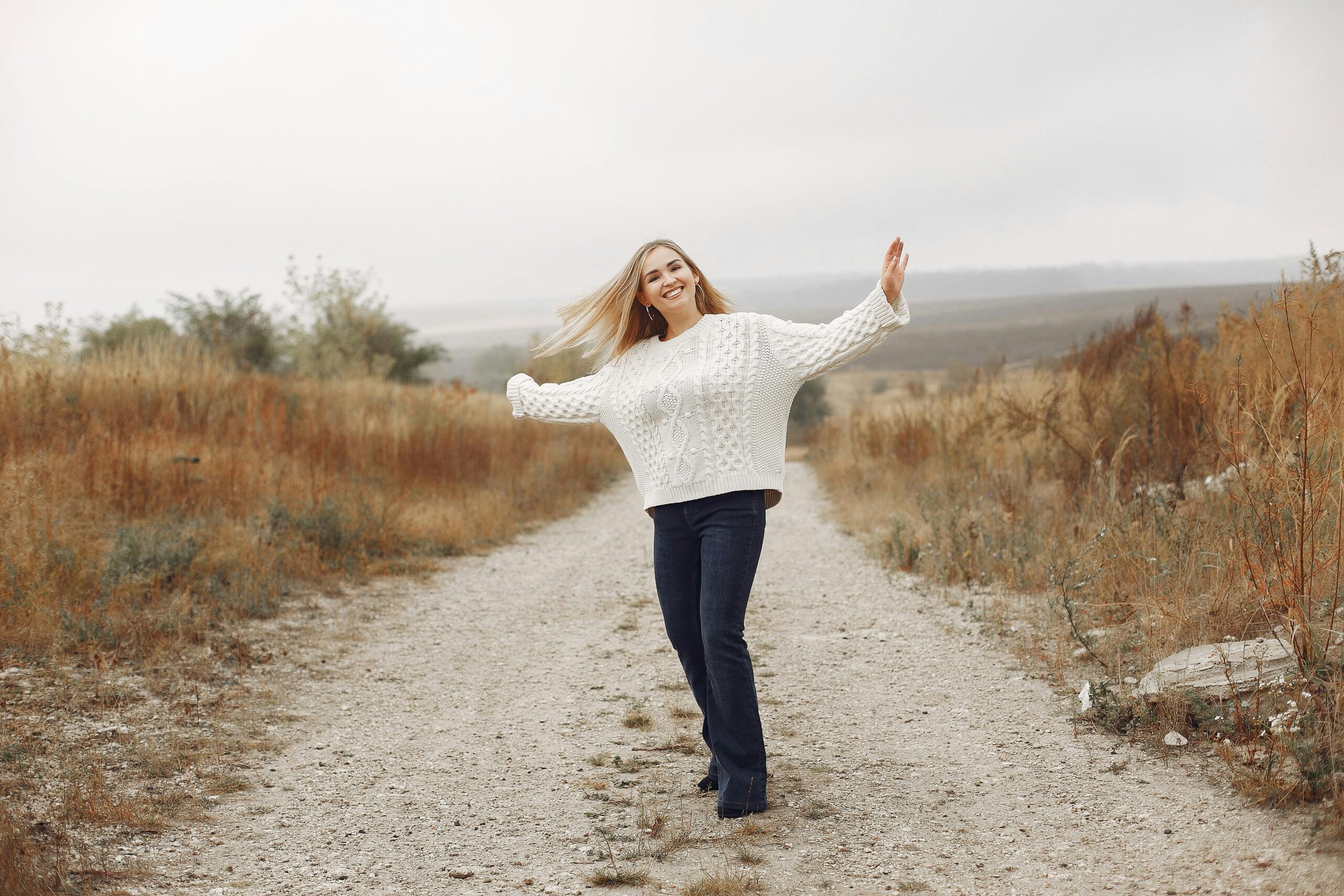 Full body of cheerful young lady in casual outfit smiling and dancing on empty rural pathway