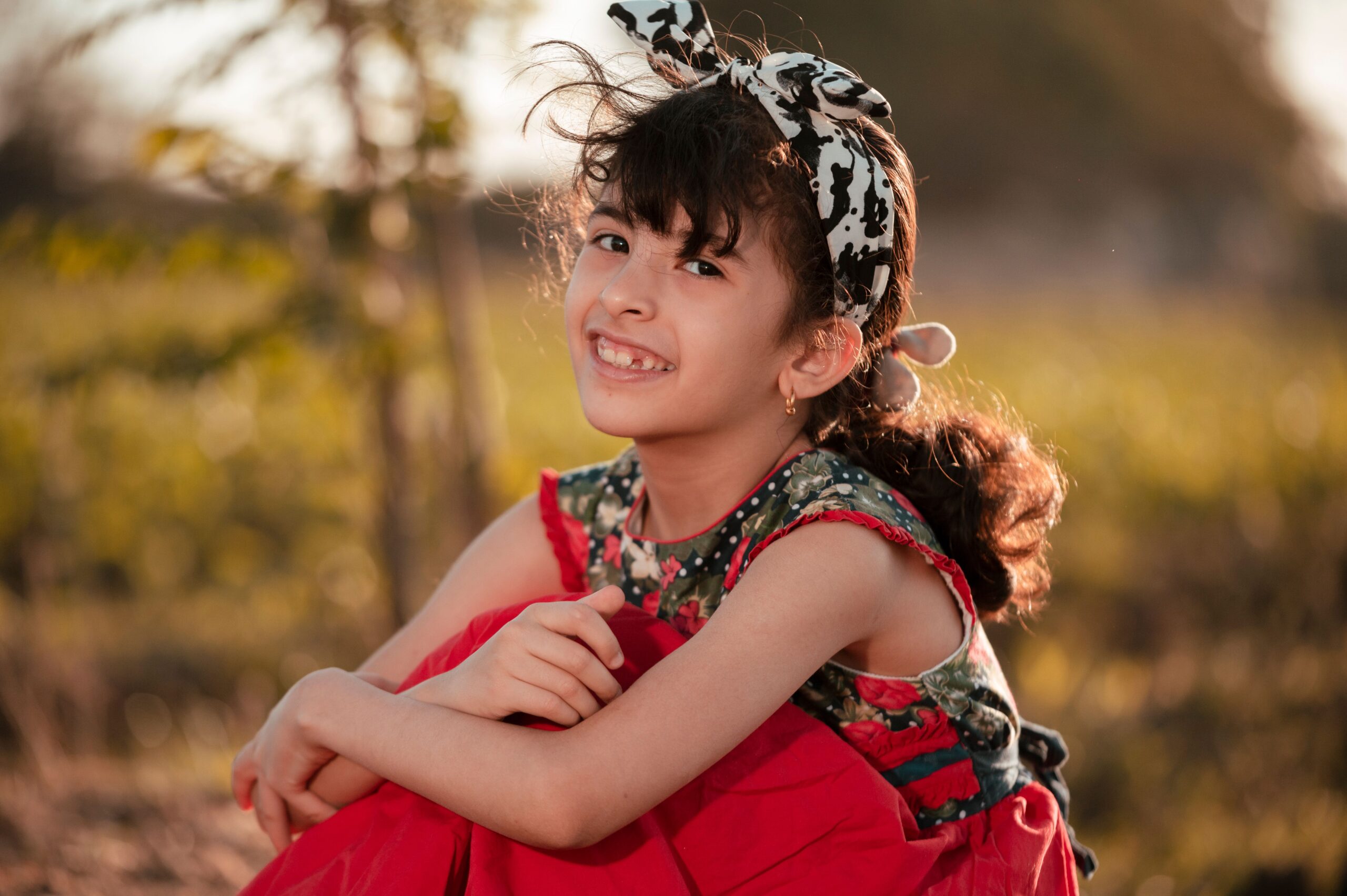 A young girl sits outdoors, smiling brightly in a red dress and headband. Captivating and cheerful.